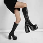 On voit les jambes d'une femme portant des bottes à talon avec une plateforme ainsi qu'une robe noire. On voit seulement ses jambes et un fond blanc.