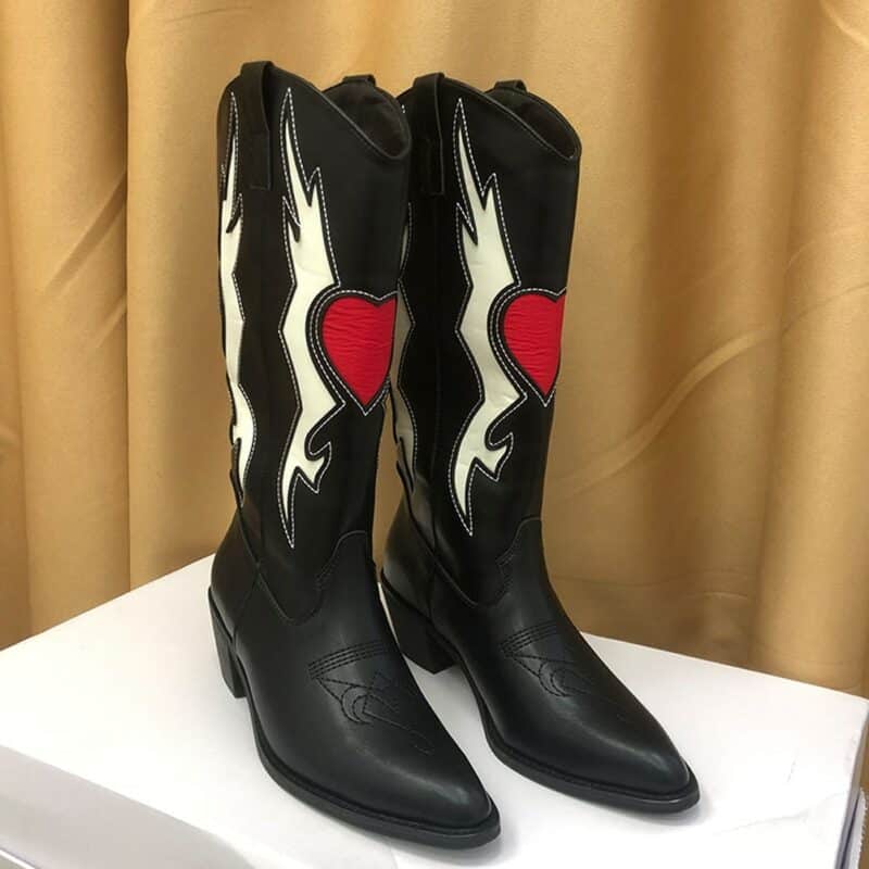Photo d'une paire de bottes de cowboy noires avec un motif de coeur ailé posé sur un cube blanc devant un rideau moutarde.