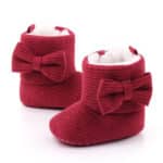 On voit deux jolies bottes en tissu, des chaussons, pour enfant, rouges avec un grand nœud sur le côté.
