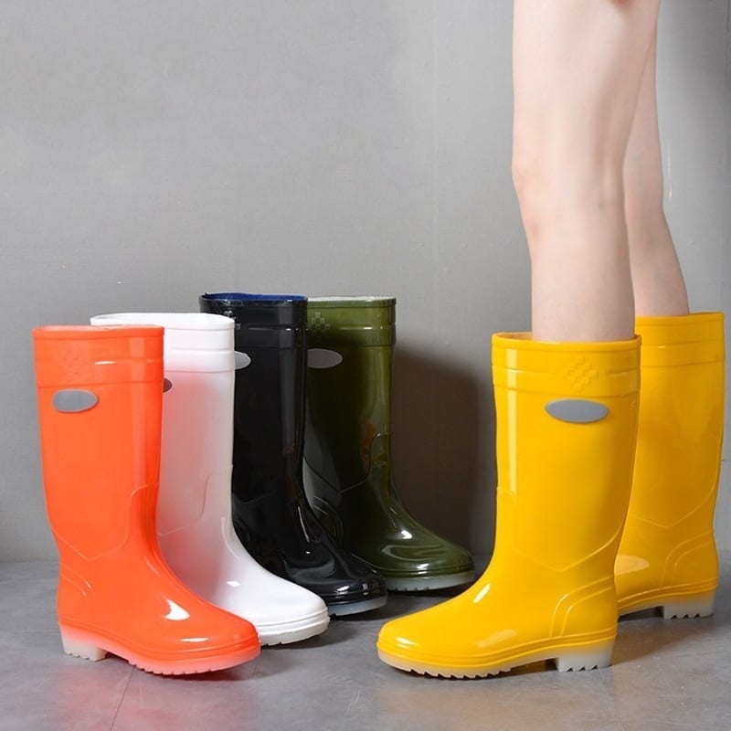 Quelqu'un dont on ne voit que les jambes, porte une paire de botte de pluie en caoutchouc jaune, et en face sont posées d'autres paires du même modèle en orange, blanc et noir