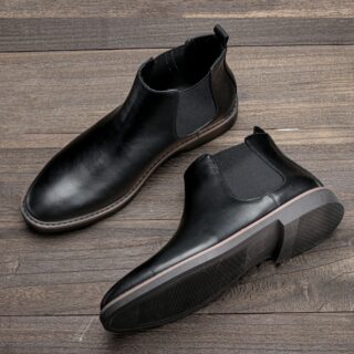 Paire de bottines noires pour homme, à enfiler posées sur un sol marron. Vu de haut, une chaussure et posée sur la semelle et l'autre à côté est posé sur le côté, avec la semelle vers le bas de la photo.
